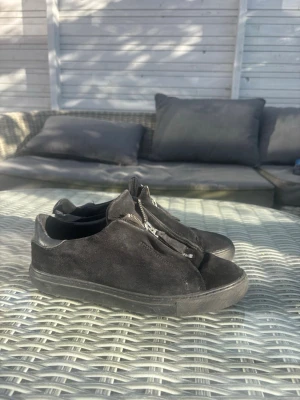 Svarta sneakers med dragkedja - Säljer ett par svarta sneakers i mocka med dragkedja framtill istället för klassiska snören. Skorna har låg profil och svart sula, samt detaljer i läder vid hälen. Perfekta för en clean och stilren look.
