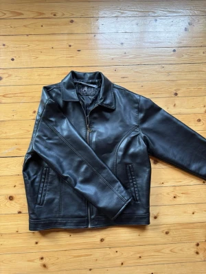 skinnjacka med dragkedja - Snygg svart skinnjacka med rak passform och dragkedja framtill. Jackan har två fickor med dragkedja och en klassisk krage. Perfekt för dig som vill ha en stilren och tidlös look. 