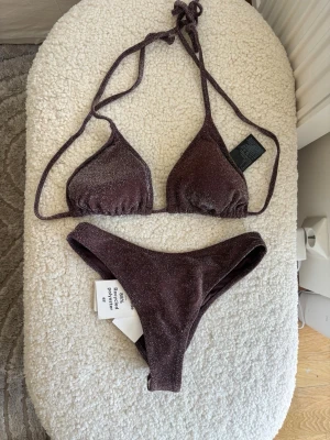 Glittrig brun bikini med knyt - Snygg brun bikini med glittrigt tyg och knyt i ryggen och nacken. Klassisk trekantsmodell upptill och matchande bikinitrosa. Perfekt för strandhäng eller poolparty. Helt oanvänd. Xs i Trosa och S i bh. Perforering kvar på trosan.