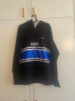 Svart hoodie från Lacoste med blå detalj - Säljer en svart hoodie från Lacoste med en bred blå rand och vita vågiga linjer över bröstet. Klassisk huva och dragsko, samt Lacoste-logga på bröstet. Hoodien har en avslappnad passform och är tillverkad i mjukt material.