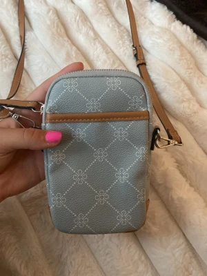 Ljusblå mönstrad crossbody-väska - Säljer en ljusblå liten handväska med vitt mönster och bruna detaljer. Väskan har justerbar axelrem, dragkedja upptill och praktiska fack både fram och bak. Perfekt storlek för mobilen och småsaker. Helt ovanvänd!  Köpt för 395kr
