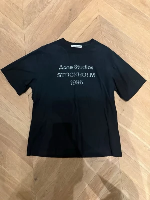 Acne Studios t-shirt - Säljer denna populära och riktigt feta t-shirt från Acne Studios med vit text 'Acne Studios STOCKHOLM 1996' på bröstet. Den är i färgen svart. Storlek S, men passar M. Klassisk passform med rund hals och korta ärmar. Helt ny, aldrig använd. Perfekt för en stilren och trendig look. Säljer den då den inte passar mig. Pris kan diskuteras