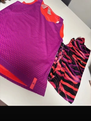 Lila träningslinne och byxor - Säljer ett lila och orange träningslinne från Adidas i meshmaterial samt ett par färgglada byxor med svart, rött och lila mönster. Perfekt för gymmet eller löpning, båda plaggen är lätta och andas bra.