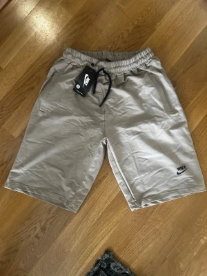 Beige shorts från Nike med snörning - Snygga beige shorts från Nike med elastisk midja och svart snörning. Klassisk Nike-logga broderad i svart på ena benet. Två sidofickor och avslappnad passform, perfekta för sommardagar eller träning.