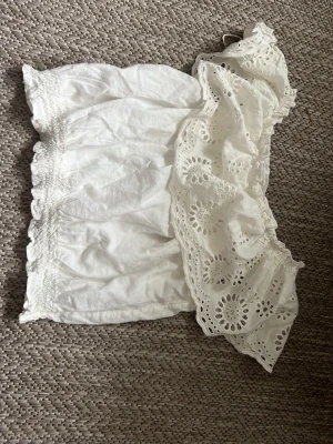 Vit offshoulder topp med spetsvolang - Söt vit offshoulder topp med bred, broderad spetsvolang och smock i nederkant. Luftig och perfekt för varma dagar, med romantisk känsla och fina detaljer. Passar dig som gillar en feminin och trendig stil.