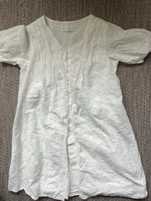 Vit broderad blus med puffärm - Söt vit blus med broderade blommönster och knappar framtill. Blusen har puffiga korta ärmar och v-ringning. Tillverkad i ett lätt tyg som känns luftigt och somrigt. Perfekt för dig som gillar romantisk och bohemisk stil.