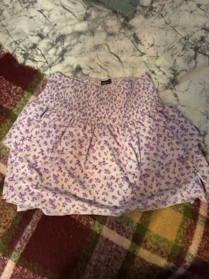 Ljuslila blommig volangkjol - Supersöt kort kjol i ljuslila med små blommor och volangdetaljer. Kjolen har bred resår i midjan för skön passform och är perfekt för vår och sommar. Materialet känns lätt och luftigt.