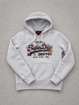 Ljusgrå hoodie från Superdry - Snygg ljusgrå hoodie från Superdry med blommigt tryck och logga på bröstet. Klassisk huva med dragsko och magficka framtill. Perfekt för en chill och avslappnad stil. Mjuk bomullsblandning och ribbade muddar vid ärmar och nederkant.