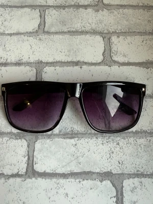 Rayband liknande glasögon - Snygga solglasögon med svart, fyrkantig båge och lila tonade glas. Klassisk modell som ger en cool vibe till din outfit. Bågarna är i plast och glasen har en lätt övergång från mörkt till ljusare lila.