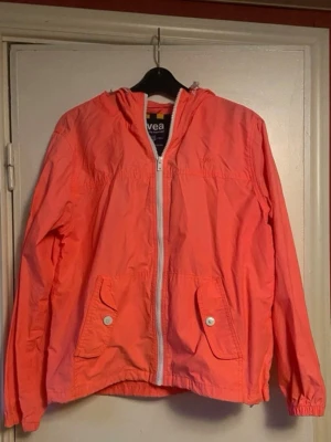 Koralrosa vindjacka med huva - Snygg korallrosa vindjacka med dragkedja framtill och två fickor med lock och knapp. Jackan har huva och resår vid ärmsluten. Perfekt för blåsiga dagar och enkel att matcha till olika outfits.rosa/orange i färgen 