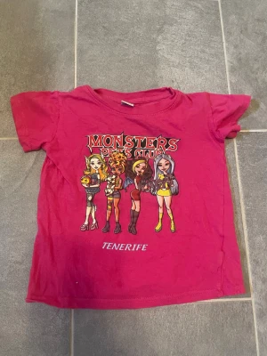 Rosa Monsters Girls Club t-shirt - Cool rosa t-shirt med tryck av Monsters Girls Club och texten Tenerife. T-shirten har korta ärmar och färgglad illustration framtill med tre tecknade figurer. Perfekt för dig som gillar färg och roliga motiv.