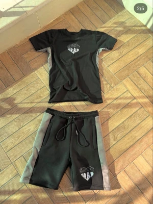 Svarta shorts med gråa detaljer - Säljer ett par svarta shorts med gråa sidopaneler och snörning i midjan. Shortsen har tryckt text på ena benet och är perfekta för en avslappnad stil. Matchar med t-shirt i samma färg och design.