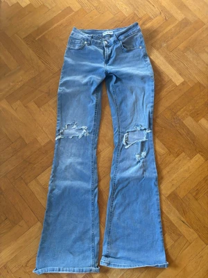 Blå slitna bootcut jeans - Säljer ett par ljusblå bootcut jeans med slitningar och hål på båda knäna. Modellen heter lowwaist men dom sitter medium waist på mig.Jeansen har klassisk femficksdesign, bälteshällor och dragkedjegylf. Perfekta för en avslappnad och trendig look.