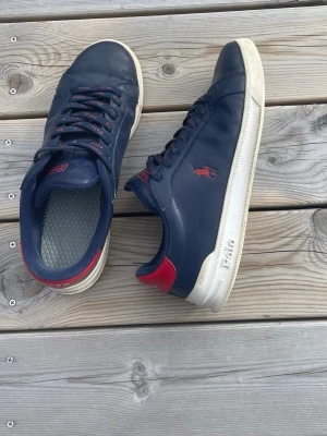 Marinblå Polo ralph lauren sneakers med röda detaljer - Snygga marinblå sneakers från Polo med röda detaljer på häl och logga. Skorna har vita sulor och klassisk låg profil. Tillverkade i slätt syntetmaterial med matchande blå och röda skosnören. Perfekta för en stilren och sportig look. Säljs dock utan sulor därav priset 