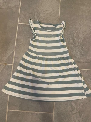 Randig kort klänning från H&M - Söt kort klänning från H&M i vitt och grönt med breda horisontella ränder. Klänningen har ärmlös design med volangdetaljer vid axlarna och är tillverkad i mjuk ekologisk bomull. Perfekt för varma dagar.