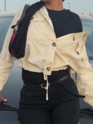 Beige cropped jeansjacka - Snygg beige cropped jeansjacka med stora knappar och dragsko i midjan. Jackan har en avslappnad passform och långa ärmar, perfekt att slänga över en t-shirt för en cool och trendig look. Från berskha