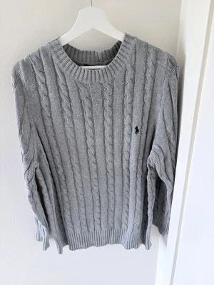 Grå kabelstickad tröja Ralph Lauren - Kabelstickad grå tröja från Ralph Lauren med rund halsringning och broderad logga på bröstet. Tröjan har långa ärmar och ribbade muddar vid ärmslut och nederkant. Perfekt för en stilren och klassisk look.