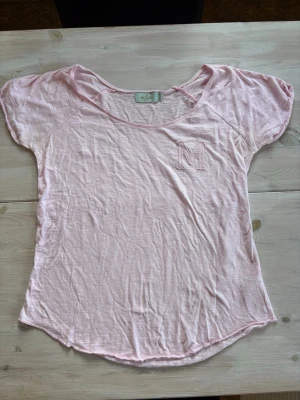 Ljusrosa t-shirt från Morris Lady - Söt ljusrosa t-shirt från Morris Lady med korta ärmar och ett broderat M på bröstet. Tillverkad i mjuk bomull och har en avslappnad passform med rund halsringning. Perfekt för en enkel och stilren look.