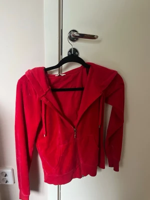 Röd velour hoodie med dragkedja - Snygg röd hoodie i mjuk velour med dragkedja framtill och två fickor. Tröjan har huva med snören och ribbade muddar vid ärmslut och nederkant. Perfekt för en chill och avslappnad stil.