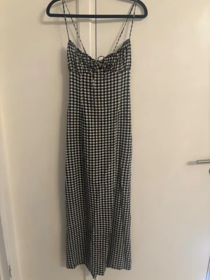 Svartvit rutig långklänning Zara  - Långklänning med smala axelband och svartvitt rutigt mönster. Klänningen har snörning framtill och är tillverkad i ett lätt material, perfekt för varma dagar. Passformen är rak och avslappnad med en feminin touch.