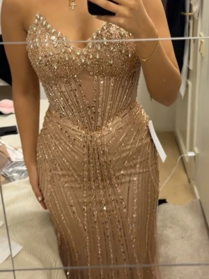 Glittrig beige långklänning - Supersnygg beige långklänning med glittriga paljettdetaljer och markerad midja. Klänningen har en djup v-ringning och figurnära passform som framhäver siluetten. Perfekt för bal eller festliga tillfällen där du vill sticka ut.