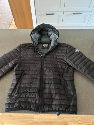 Svart dunjacka med huva från Colmar - Snygg svart dunjacka från Colmar med quiltad design och fast huva. Jackan har två dragkedjeförsedda fickor framtill och Colmar-logga på ärmen. Insidan av huvan är ljusgrå. Perfekt för kalla vinterdagar.