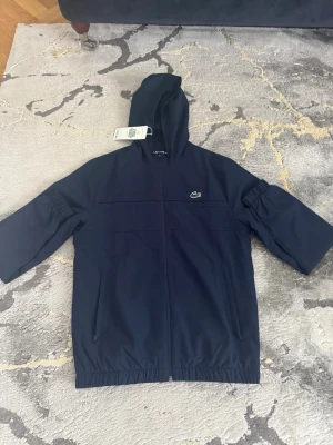Mörkblå vindjacka från Lacoste ny pris 1350kr - Helt ny Snygg mörkblå vindjacka från Lacoste med huva och dragkedja framtill. Jackan har det klassiska gröna krokodilmärket på bröstet, två sidofickor och elastisk nederkant. Materialet är tunt och passar perfekt för blåsiga dagar.