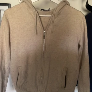 Beige hoodie med dragkedja - Mysig beige hoodie med dragkedja framtill och två fickor. Tillverkad i ett mjukt, stickat material och har en klassisk huva med dragsko. Perfekt för en avslappnad och chill stil.