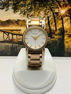Guldig klocka från DKNY med stenar - Snygg guldig klocka från DKNY med länkarmband i metall och en rund urtavla. Urtavlan är vit med glittriga stenar som timmarkeringar och en ram med små stenar runt. Perfekt accessoar för att lyfta din stil.