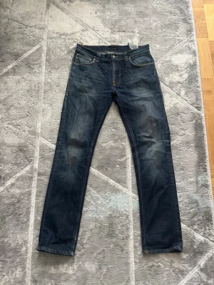 Mörkblå nudie jeans med passformen slim fit  - Säljer ett par mörkblå jeans med modellen slim fit och klassisk femficksmodell. Jeansen har kontrastsömmar och slitningar på både fram- och baksida samt detaljerade bakfickor med broderad design. Perfekta för en avslappnad och trendig look.