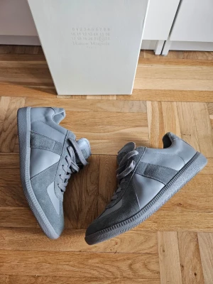 Maison Margiela Gats Full Grey Sneakers - Snygga grå sneakers från Maison Margiela med klassisk siluett. Skorna har en mix av mocka och slätt läder, tonade grå nyanser och matchande grå snörning. Perfekta för dig som gillar stilrena och minimalistiska sneakers med premiumkänsla. Med box osv