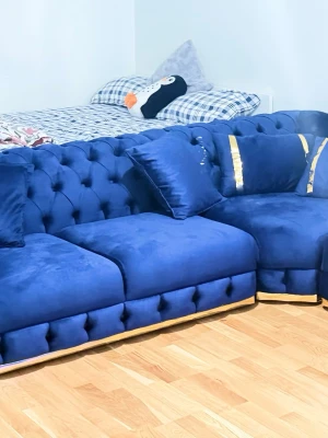 Blå sammetssoffa med guld-detaljer - Stilren hörnsoffa i djupblå sammet med tuftade detaljer och dekorativa guldfärgade band längst ner och på kuddarna. Soffan har flera matchande kuddar och en lyxig känsla som passar perfekt i vardagsrummet.