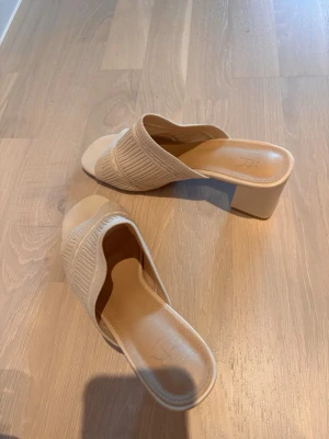 Beige sandaler med klack - Säljer ett par snygga beige sandaler med bred stickad ovandel och fyrkantig öppen tå. Skorna har en stabil blockklack och en mjuk innersula för extra komfort. Perfekta till sommarens outfits och lätta att matcha med det mesta.
