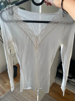 Intimissimi topp - Säljer en vit långärmad topp med vacker spets runt v-ringningen och vid ärmsluten. Toppen har en smickrande passform och är tillverkad i ett mjukt material som känns skönt mot huden. Bra att styla med jeans eller kjol för en trendig look.