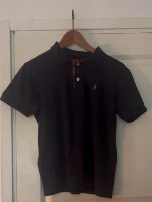 Mörkblå prickig pikéskjorta - Mörkblå pikéskjorta med små vita och röda prickar, klassisk krage och korta ärmar. Broderad brun logga på bröstet och knappar vid halsen. Skön och stilren modell.