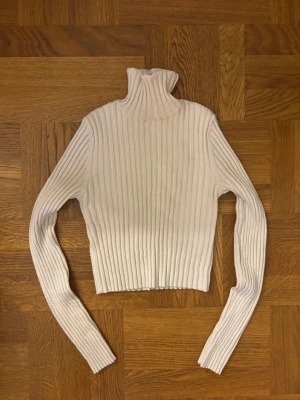 Vit ribbad cropped polotröja - Säljer en vit, ribbstickad cropped polotröja med långa ärmar. Tröjan har en tight passform och hög krage, perfekt för lager på lager och trendiga outfits. Materialet är mjukt och stretchigt, vilket gör den bekväm att bära.