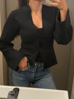 Svart kofta med vida ärmar - Stilren svart kavaj med tre knappar framtill och trendigt vida ärmar. Kavajen har en något figurnära passform och en klassisk krage utan slag. Perfekt att matcha med jeans för en cool look.