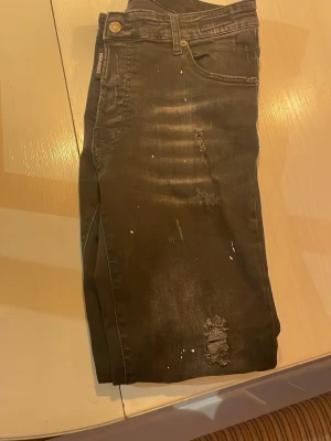 Svarta slim jeans med slitningar - Säljer ett par svarta skinny jeans med slitningar och små vita färgstänk. Jeansen har klassisk femficksmodell, låg midja och smal passform. Perfekta för en trendig och avslappnad look.
