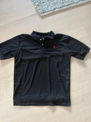 Svart pikétröja från Polo Ralph Lauren - Klassisk svart pikétröja från Polo Ralph Lauren med röd broderad logga på bröstet. Tröjan har korta ärmar, krage och knäppning med två knappar. Perfekt till både jeans och shorts för en clean och stilren look. Storlek L men passar även M.