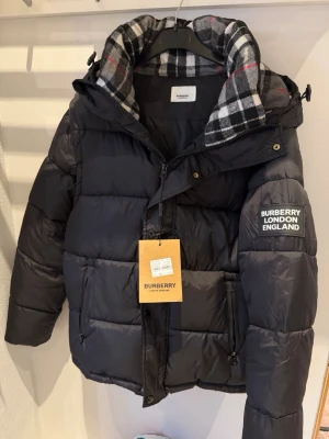 Svart dunjacka från Burberry - Snygg svart dunjacka från Burberry med klassiskt rutigt foder i huvan och logotyp på ärmen. Jackan har dragkedja, fickor med dragkedja och hög krage. Perfekt för kalla vinterdagar och riktigt trendig look.