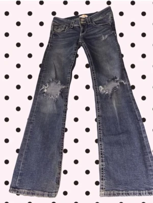 Blå bootcut jeans med slitningar - Säljer ett par blå bootcut jeans med tydliga slitningar och hål på båda benen. Jeansen har låg midja, klassiska fem fickor och vita kontrastsömmar. Perfekta för dig som gillar en avslappnad och trendig look.