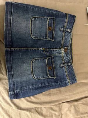 Mörkblå jeanskjol med fickor - Snygg mörkblå jeanskjol med två stora fickor framtill, dekorerade med bruna knappar. Kjolen har tydliga gula sömmar och bälteshällor i midjan. Perfekt för en avslappnad och trendig look.