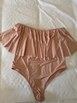Nude off shoulder body Kisinki M - Supersnygg nude-färgad body från Kisinki i off shoulder-modell med bred volang upptill. Tillverkad i mjukt och stretchigt material som sitter skönt mot kroppen. Perfekt för dig som vill ha en trendig och bekväm look.