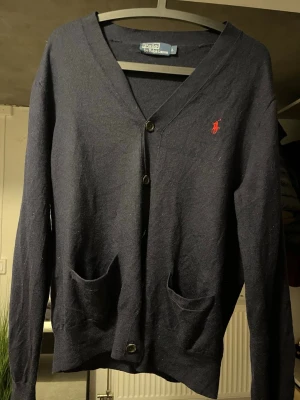 Mörkblå cardigan från Polo Ralph Lauren - Klassisk mörkblå cardigan från Polo Ralph Lauren med röd broderad logga på bröstet. Tröjan har v-ringning, knäppning framtill och två fickor. Perfekt att slänga över en t-shirt för en chill och stilren look.