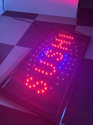 LED-skylt med röd och grön text - Elektronisk LED-skylt med lysande röda, gröna och blå dioder som bildar texten 'OPEN'. Skylten har en svart rektangulär ram och ansluts med en svart sladd. Perfekt för att visa öppet-läge i butik eller café. Kontakta mig vid intresse 
