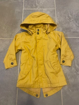 Gul parkas med huva och knappar - Snygg gul parkas med huva och knappar framtill. Jackan har två stora fickor, långa ärmar med knappdetaljer och längre bakstycke. Perfekt för dig som vill sticka ut med en färgglad och praktisk jacka i vardagen.