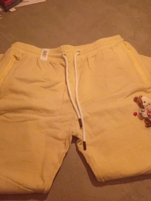 Gula mjukisbyxor med nallepatch - Säljer ett par ljusgula mjukisbyxor med vita dragsnören och dragkedjeförsedda fickor. På ena benet finns en broderad nallepatch som detalj. Byxorna är i mjukt material, perfekta för chill dagar hemma eller när du vill vara bekväm.