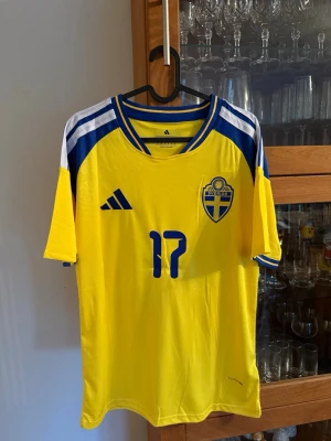 Svensk landslagströja nr 17 Gyökeres - Gul och blå svensk landslagströja från Adidas med nummer 17 och namnet Gyökeres på ryggen. Tröjan har korta ärmar, blå detaljer på axlarna och ett broderat landslagsemblem på bröstet. Perfekt för fotbollsfans som vill stötta Sverige.