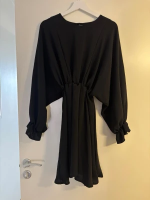 Svart klänning med volangärm - Svart kortklänning med markerad midja. Klänningen har långa, lösa ärmar med volangdetaljer vid ärmsluten och ett mjukt, flytande tyg som ger en elegant siluett. Perfekt för dig som gillar stilrena och trendiga plagg.