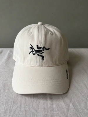 Arcteryx keps - Snygg vit keps i bomull med böjd skärm och svart broderad logga framtill. Justerbar rem baktill med metallspänne. Perfekt accessoar för en clean och stilren look.